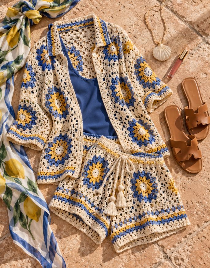Flat lay of a blue and yellow crochet shirt and shorts set over a blue tank, with a lemon-print scarf, a shell pendant, lip gloss and tan leather slides on stone