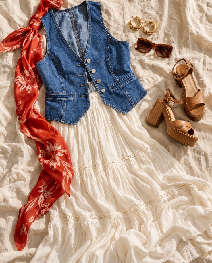 Flat lay of a fitted denim waistcoat over a tiered white maxi skirt, with a red floral scarf, gold hoops, tortoiseshell sunglasses and tan platform sandals on cream linen