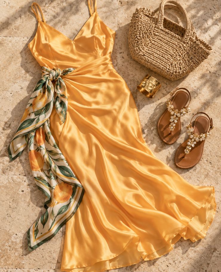 Flat lay of a lemon satin slip dress with a lemon-print silk scarf tied at the waist, a woven straw bag, a gold cuff and embellished flat sandals on warm stone