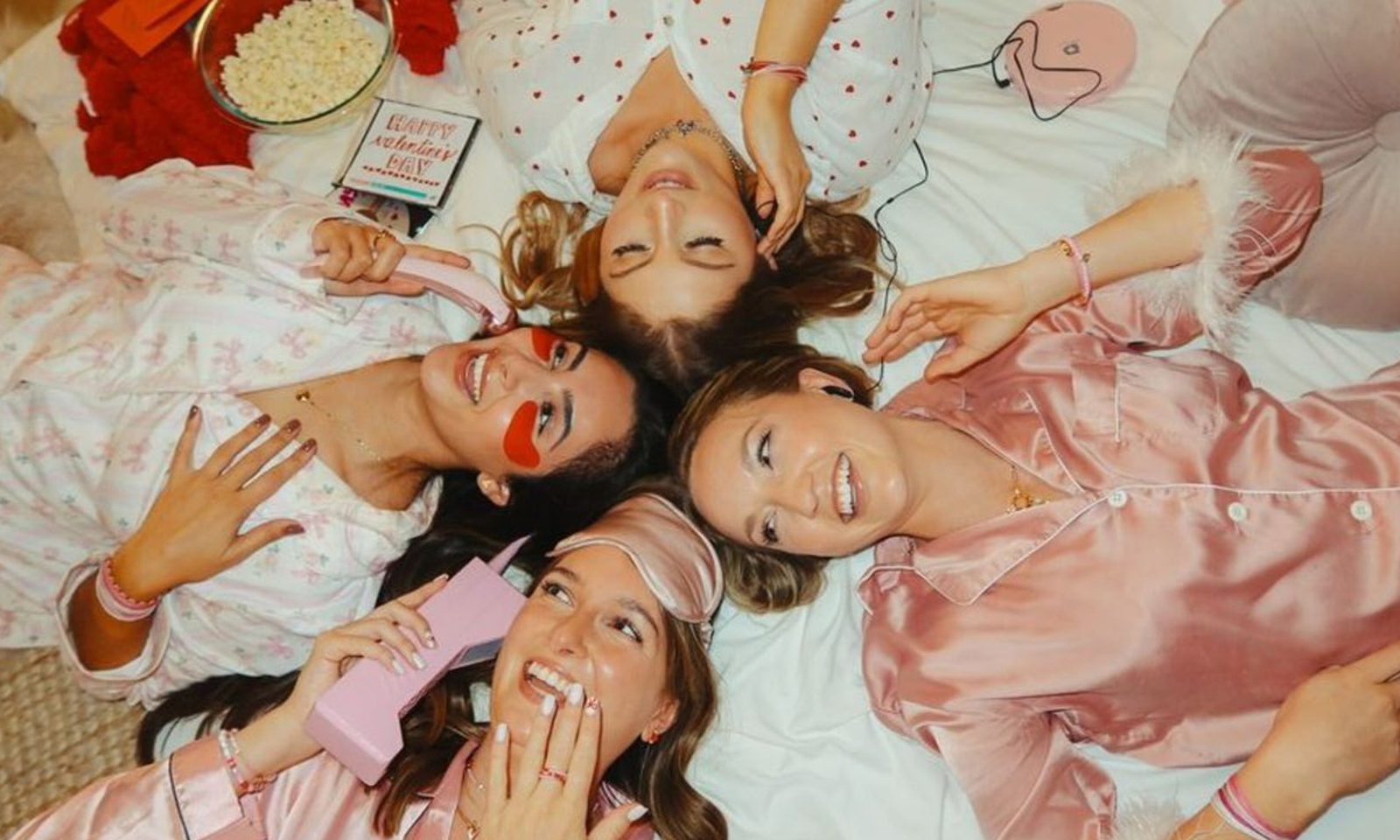 Friends in pink satin pyjamas lying head to head on a bed, laughing