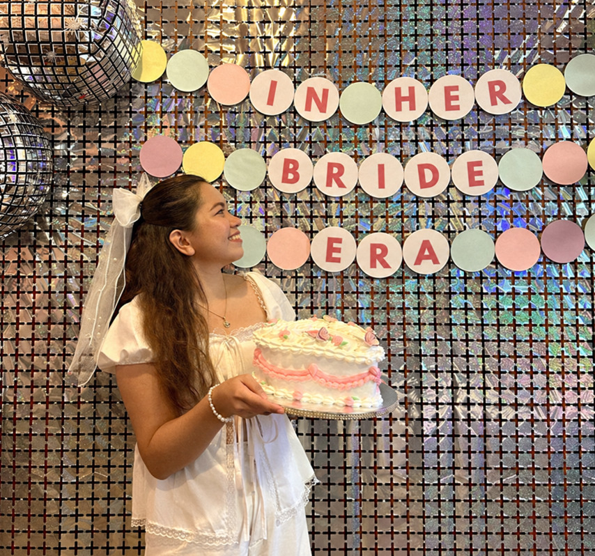 A bride-to-be in a white dress with a big white bow and veil holding a pink heart cake in front of a sequin wall
