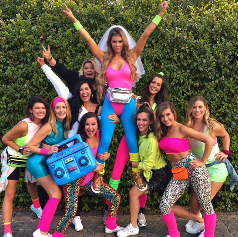 A bride-to-be in a veil, pink leotard and leg warmers lifted up by friends in neon leotards, leg warmers and bum bags, one holding a blue boombox