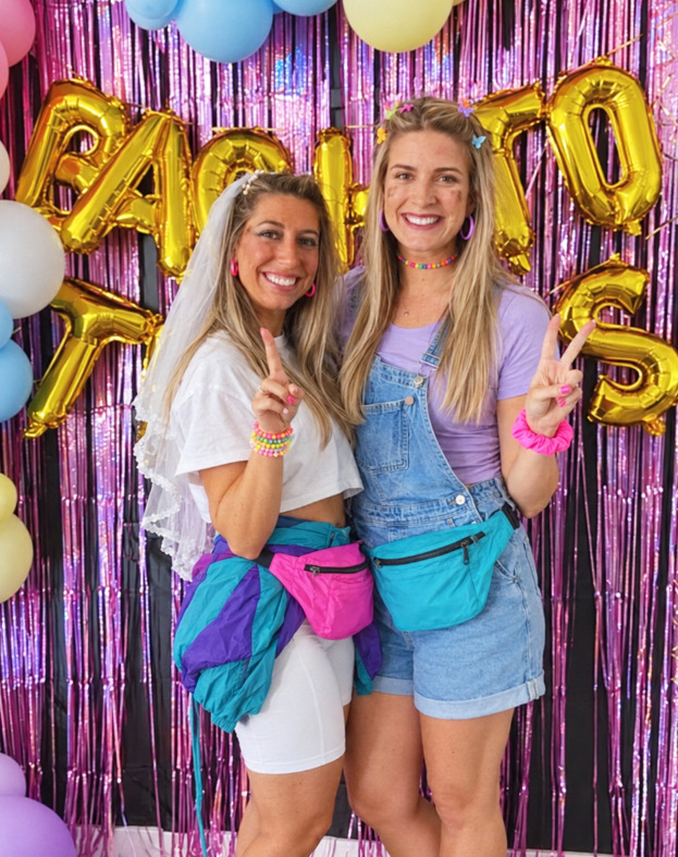 A bride-to-be in a veil, white tee and bike shorts with a windbreaker tied at her waist and a bum bag, beside a friend in denim overalls, both making peace signs under gold balloons
