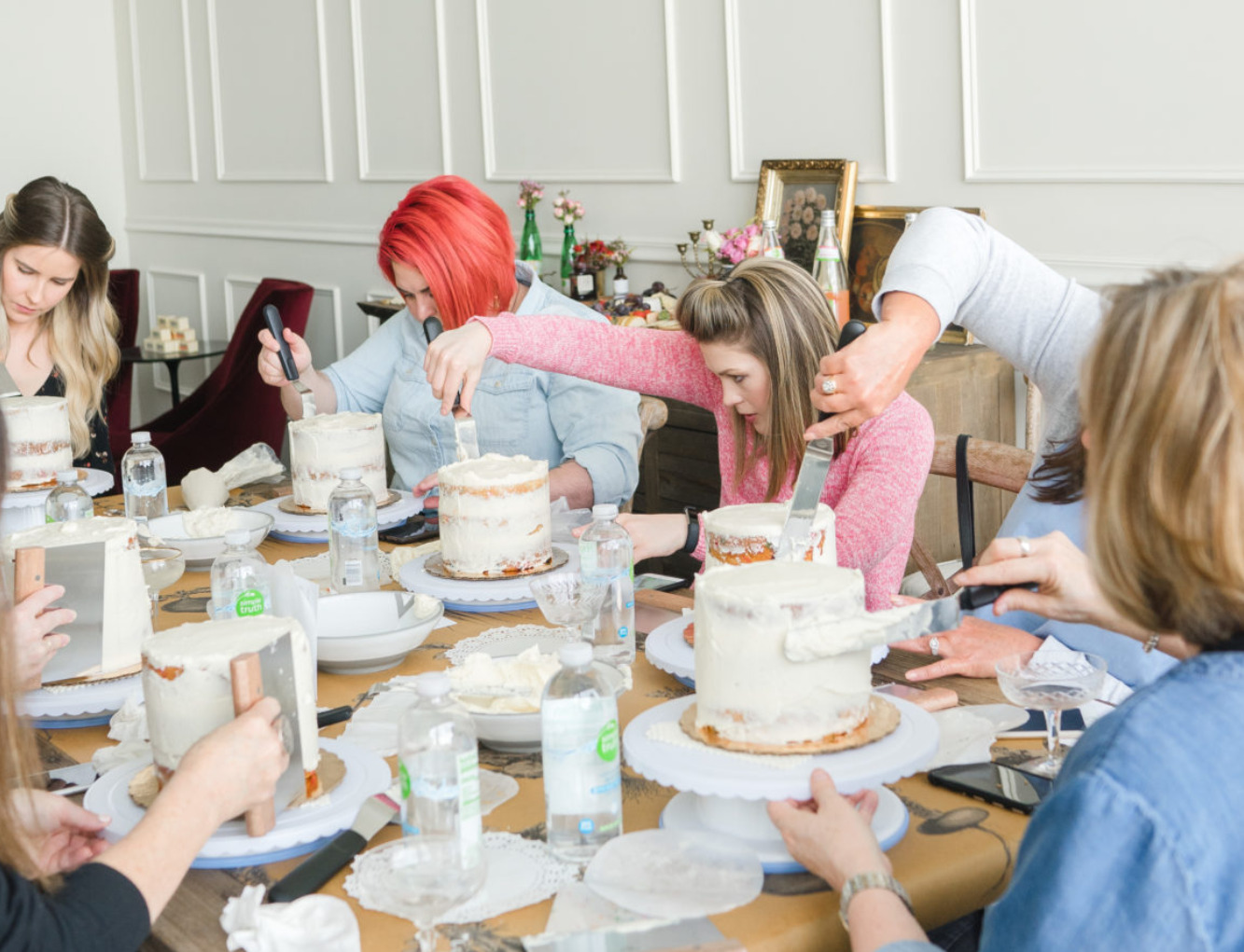 Friends at a long table icing naked cakes with spatulas on turntables