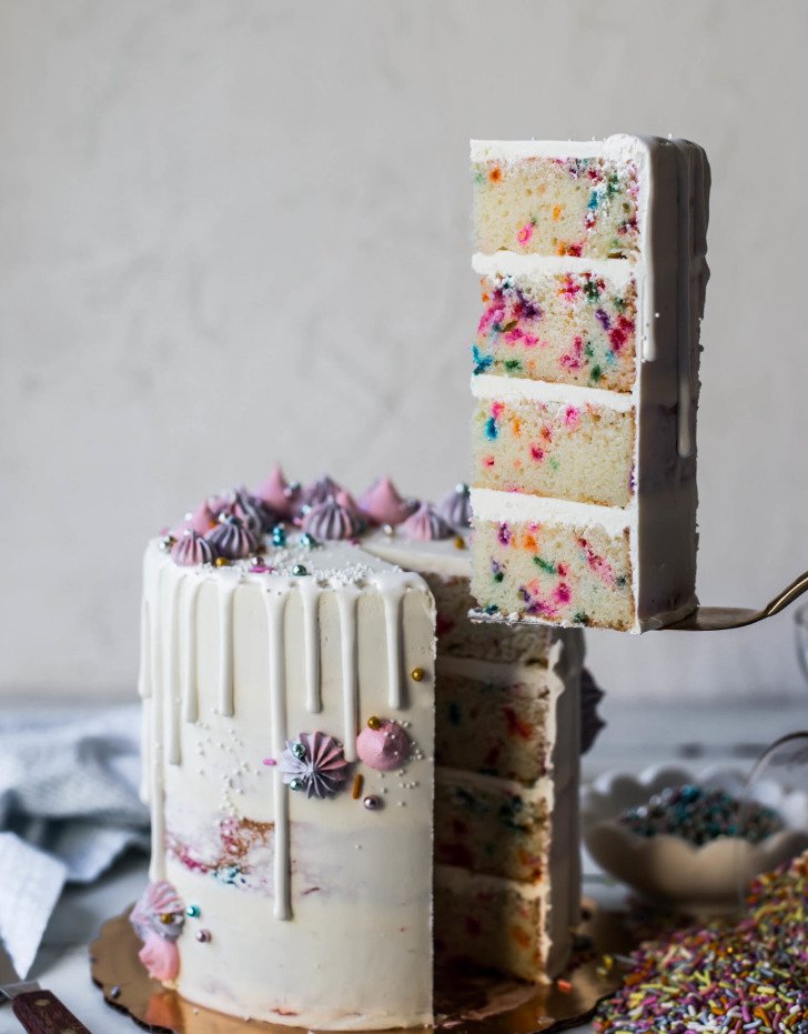 A funfetti layer cake with a slice lifted out, a white drip and pastel meringues
