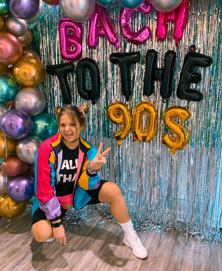 A friend in a colour-block windbreaker and white sneakers crouching with a peace sign in front of a blue tinsel curtain, chrome balloons and big black and gold letter balloons