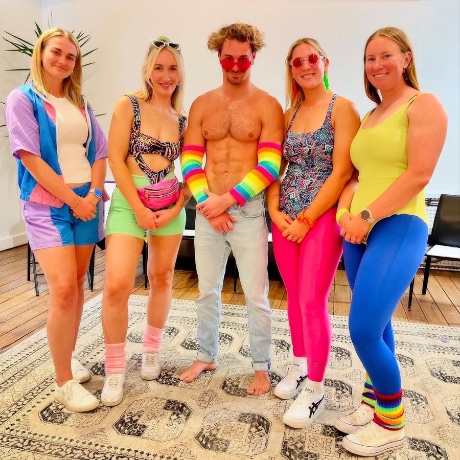 Hens in 80s aerobics outfits with a shirtless model in jeans and rainbow arm warmers