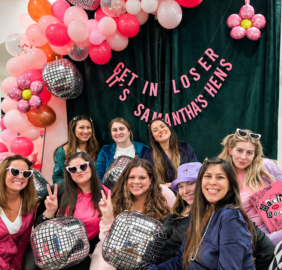 A hens group in heart sunglasses, a bucket hat and denim holding disco-ball balloons under a pink balloon arch and a GET IN LOSER banner