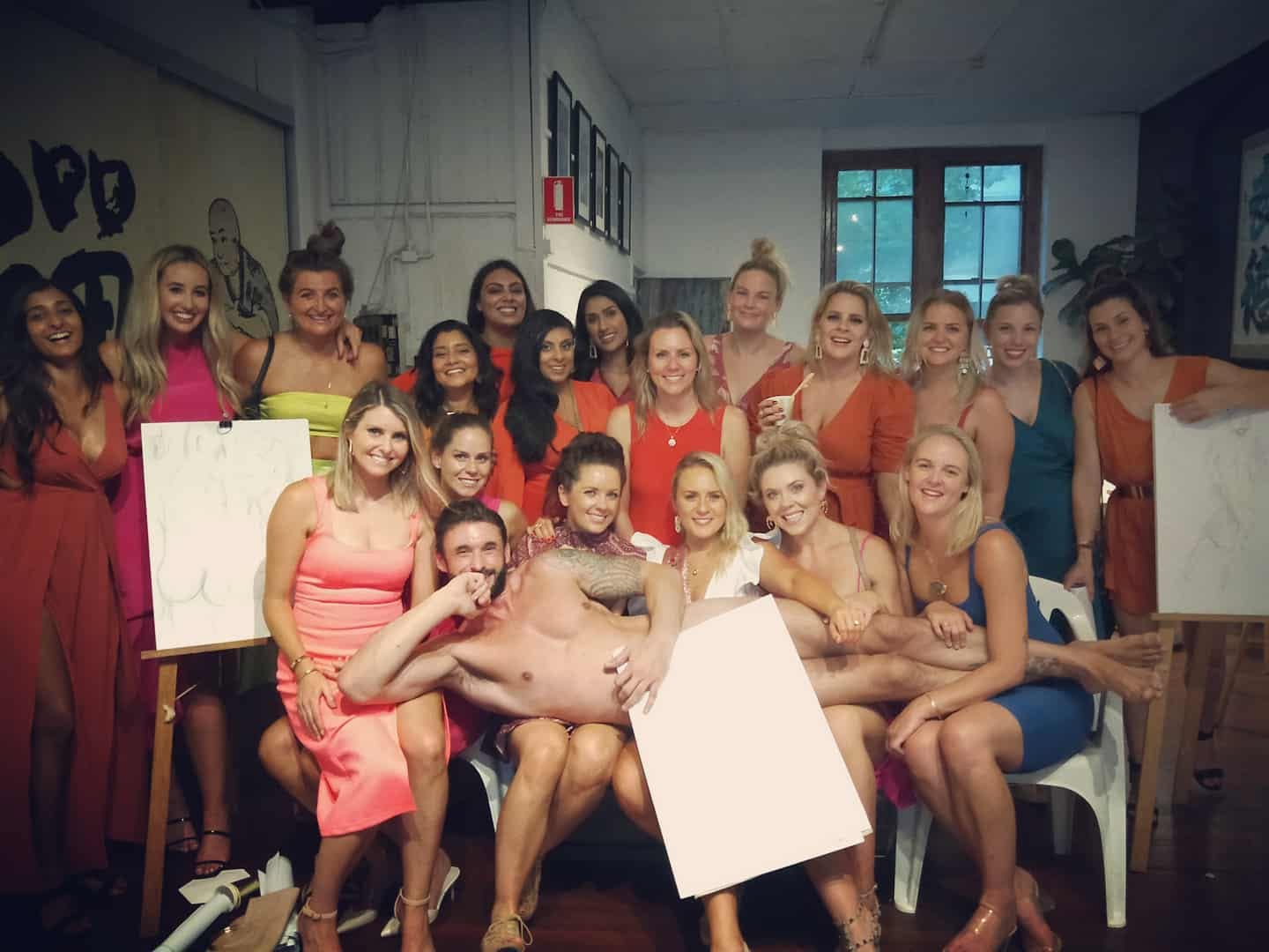 A big hens group in bright dresses laughing in a drawing studio, easels with sketches either side, the life drawing model lying across their laps behind a sheet of paper