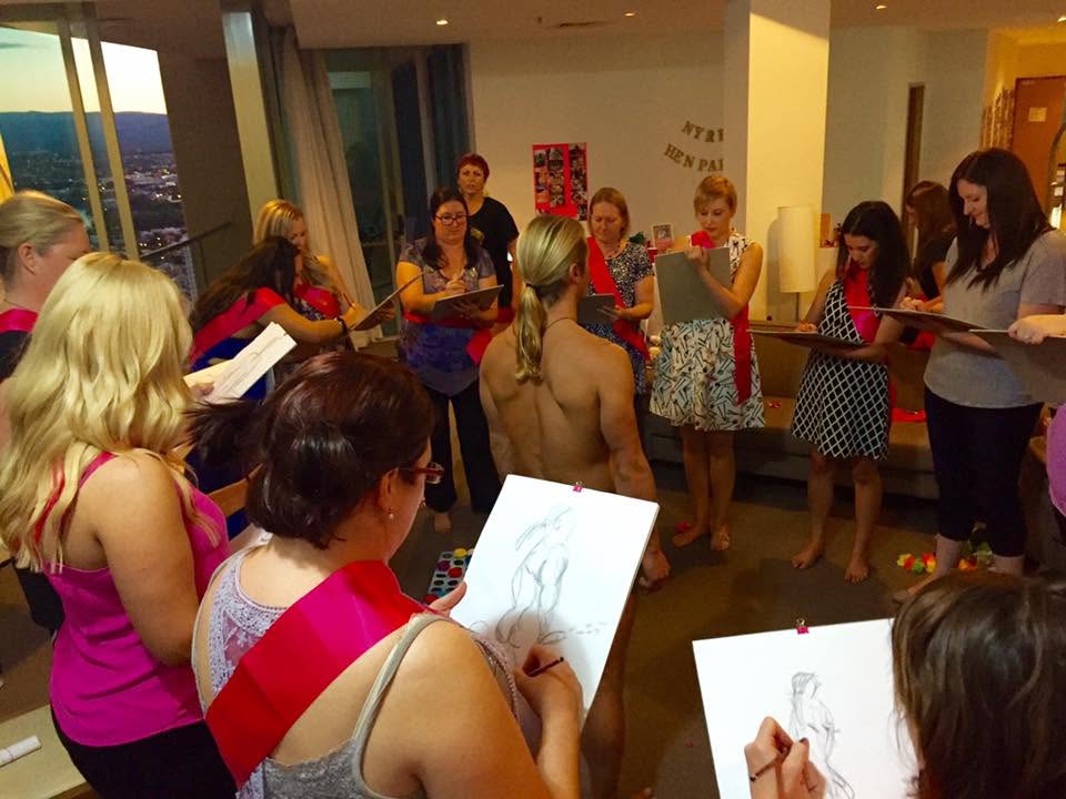 Hens in pink sashes standing in a loose circle with sketch pads, drawing the life drawing model in a lounge room