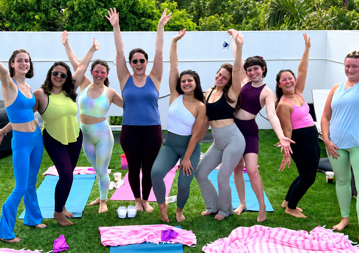 A group of friends in bright activewear with their arms in the air beside yoga mats on a sunny lawn