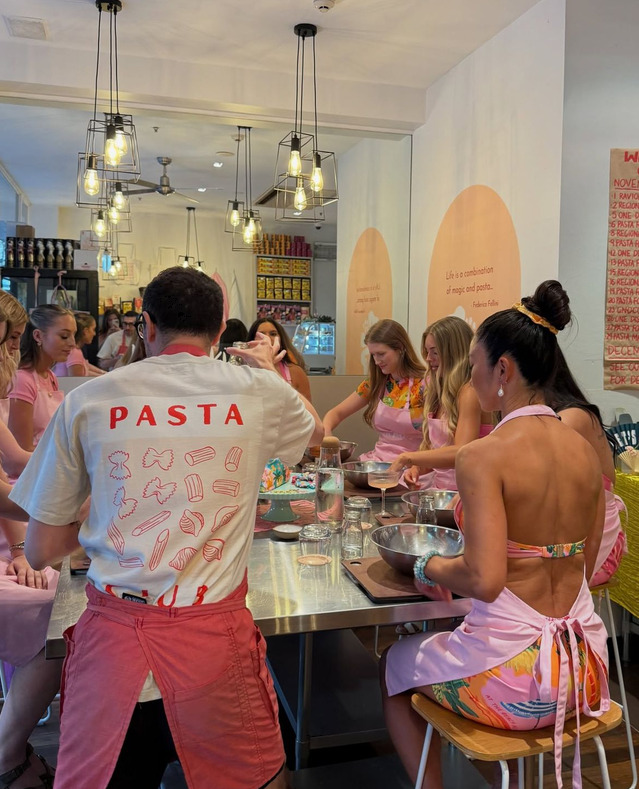 A pasta class host in a PASTA tee with women in pink aprons at a steel bench
