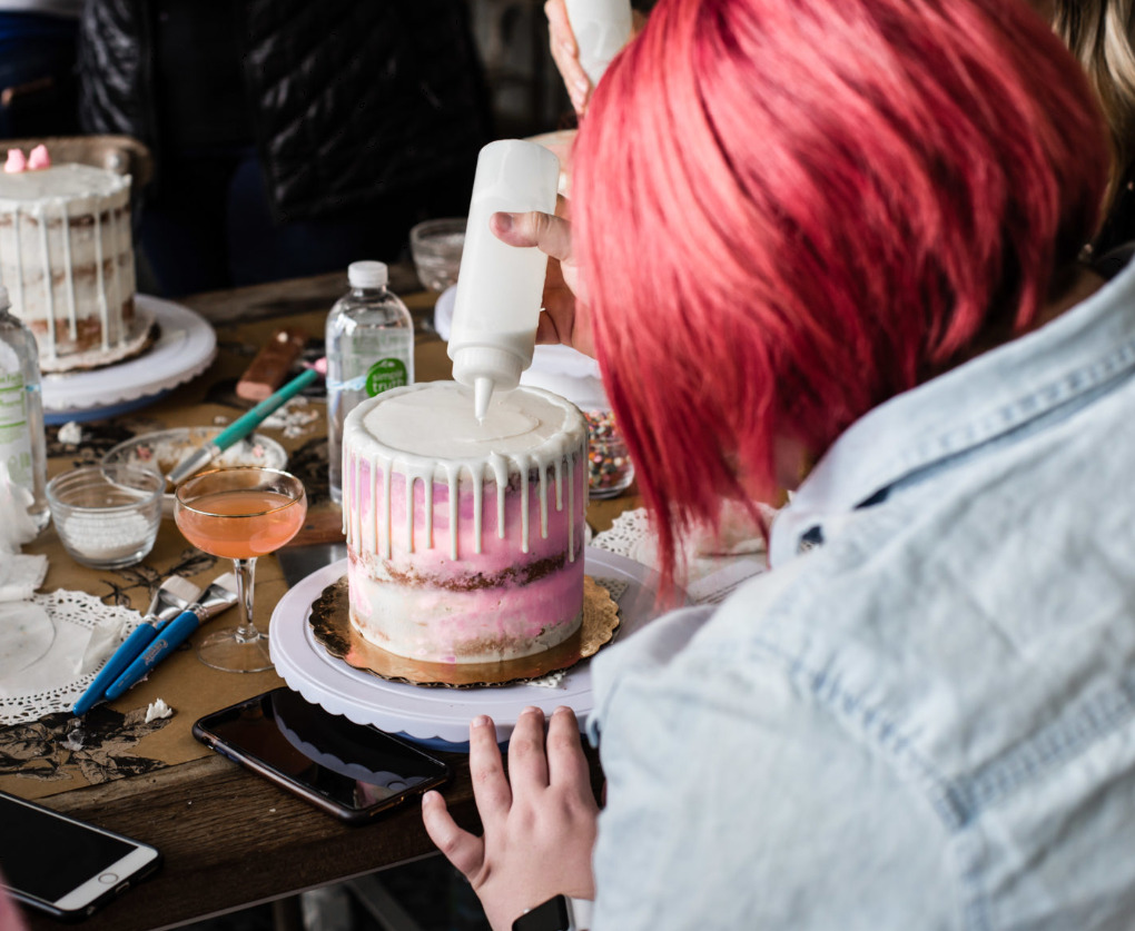 A woman with red hair drizzling white ganache over a pink drip cake