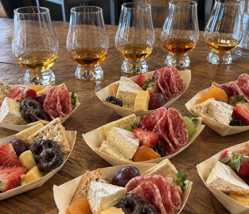 Five whisky tasting glasses behind grazing boats of cheese, salami and fruit on a timber table