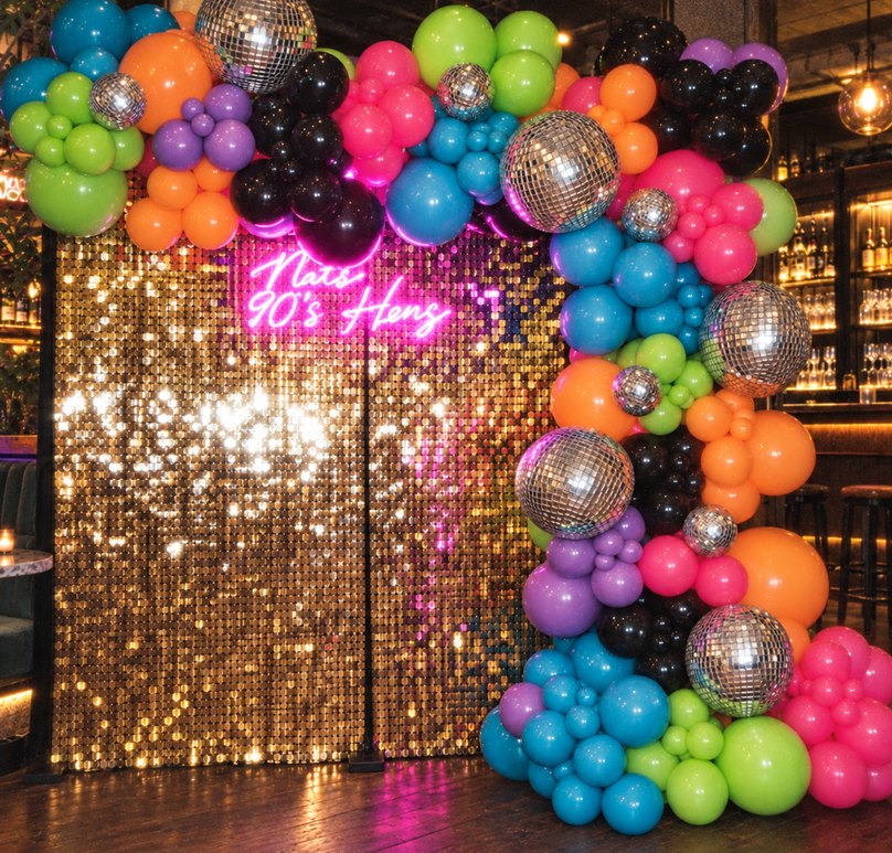 A balloon garland in hot pink, lime, orange, aqua, purple and black with mirror balls tucked through it, wrapped around a gold sequin wall with a pink neon sign reading Nat’s 90’s Hens