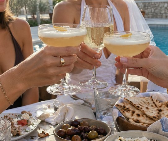 Coupes and a flute raised over olives, pita and share plates beside a pool