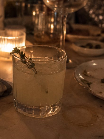 A pale lemon drink with a sprig of thyme in a tumbler beside candles