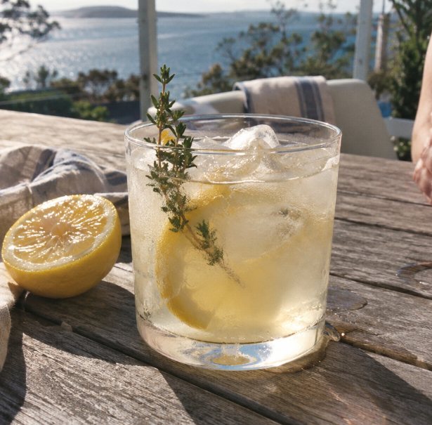 A gin and lemon cocktail with a sprig of thyme on a sunny outdoor table above the water
