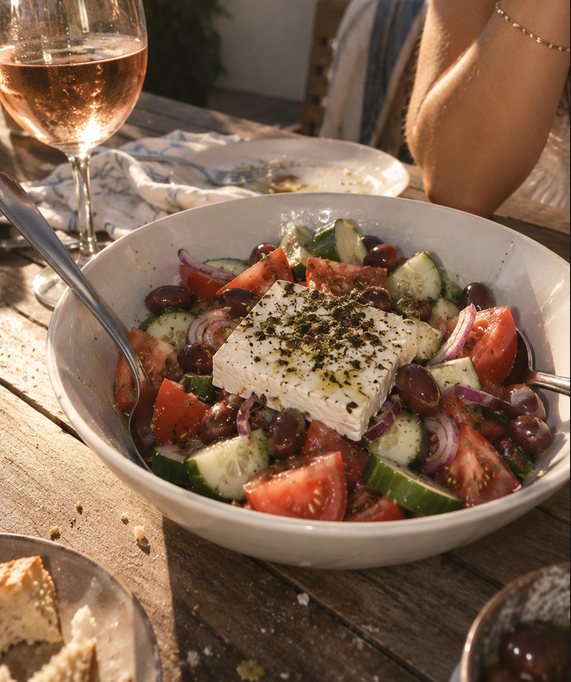 A Greek salad of tomato, cucumber, red onion and olives with a slab of feta on top, in late sun