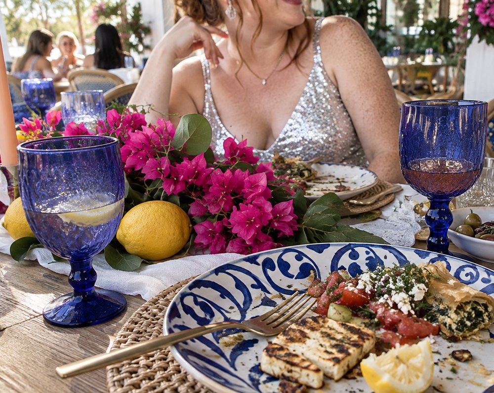 A long table set with blue goblets, lemons and bougainvillea, a plate of halloumi, Greek salad and spanakopita, a guest in silver sequins behind