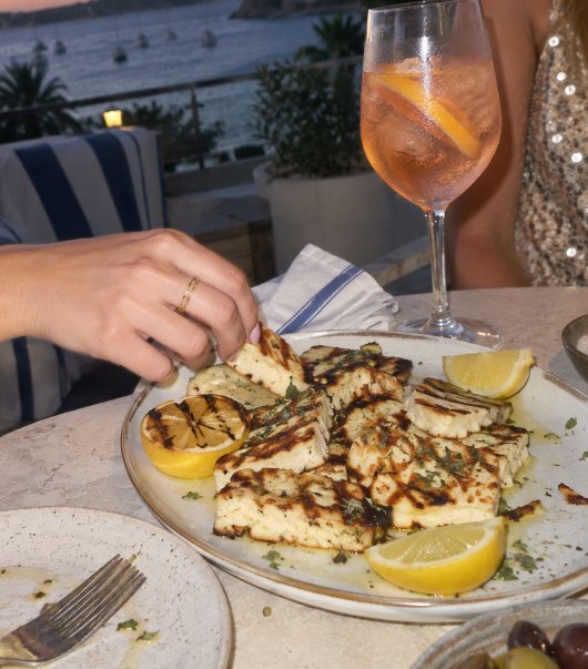 A hand reaching for grilled halloumi with lemon, a spritz and a sequin dress beside it, boats on the harbour behind