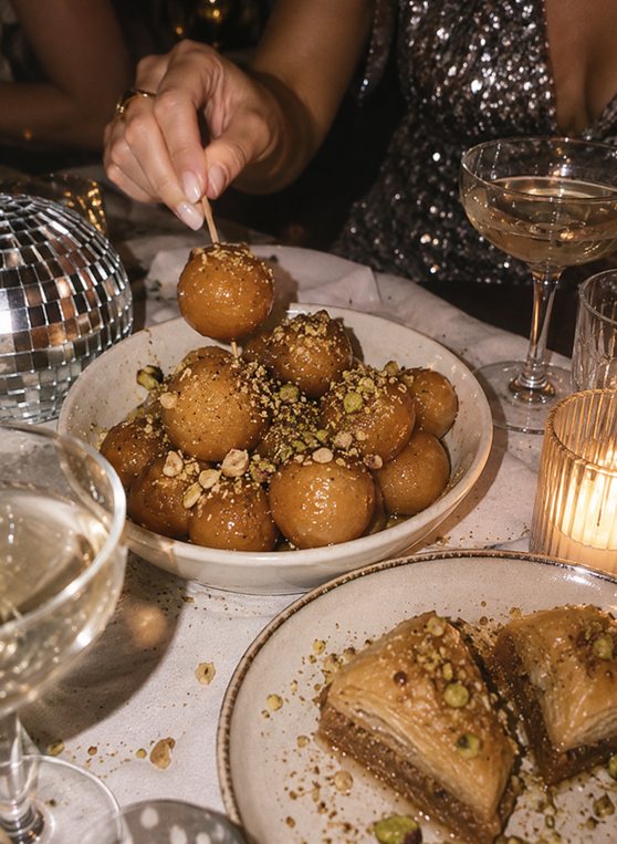Honey loukoumades with pistachio and baklava beside a mini disco ball, a sequin dress and champagne
