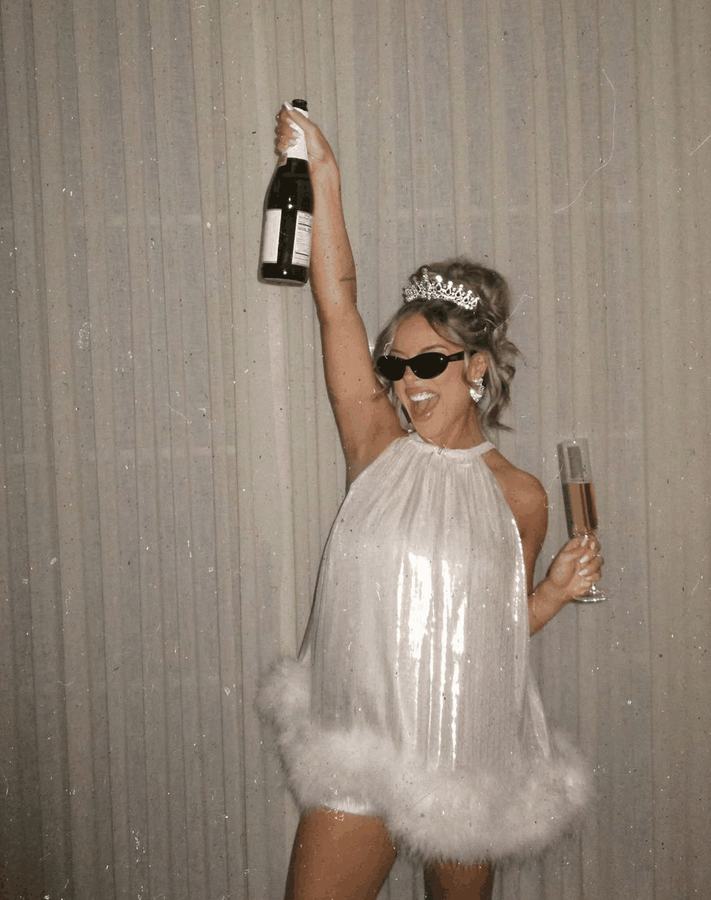 A bride to be in a feather-trimmed dress holding a champagne bottle above her head