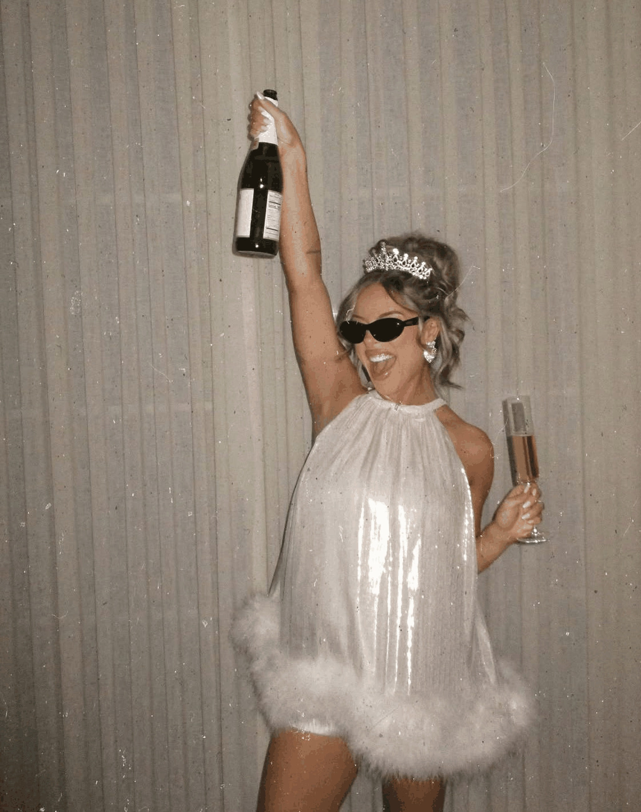 A bride to be in a tiara and a white feather-trimmed dress holding a champagne bottle above her head
