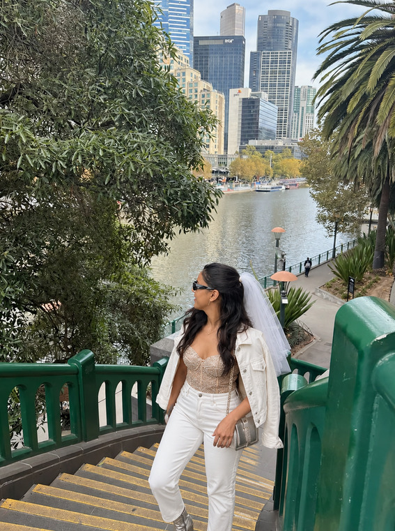 A bride to be in a veil and an all-white outfit on the riverside stairs by the Yarra, with the Melbourne city towers behind her