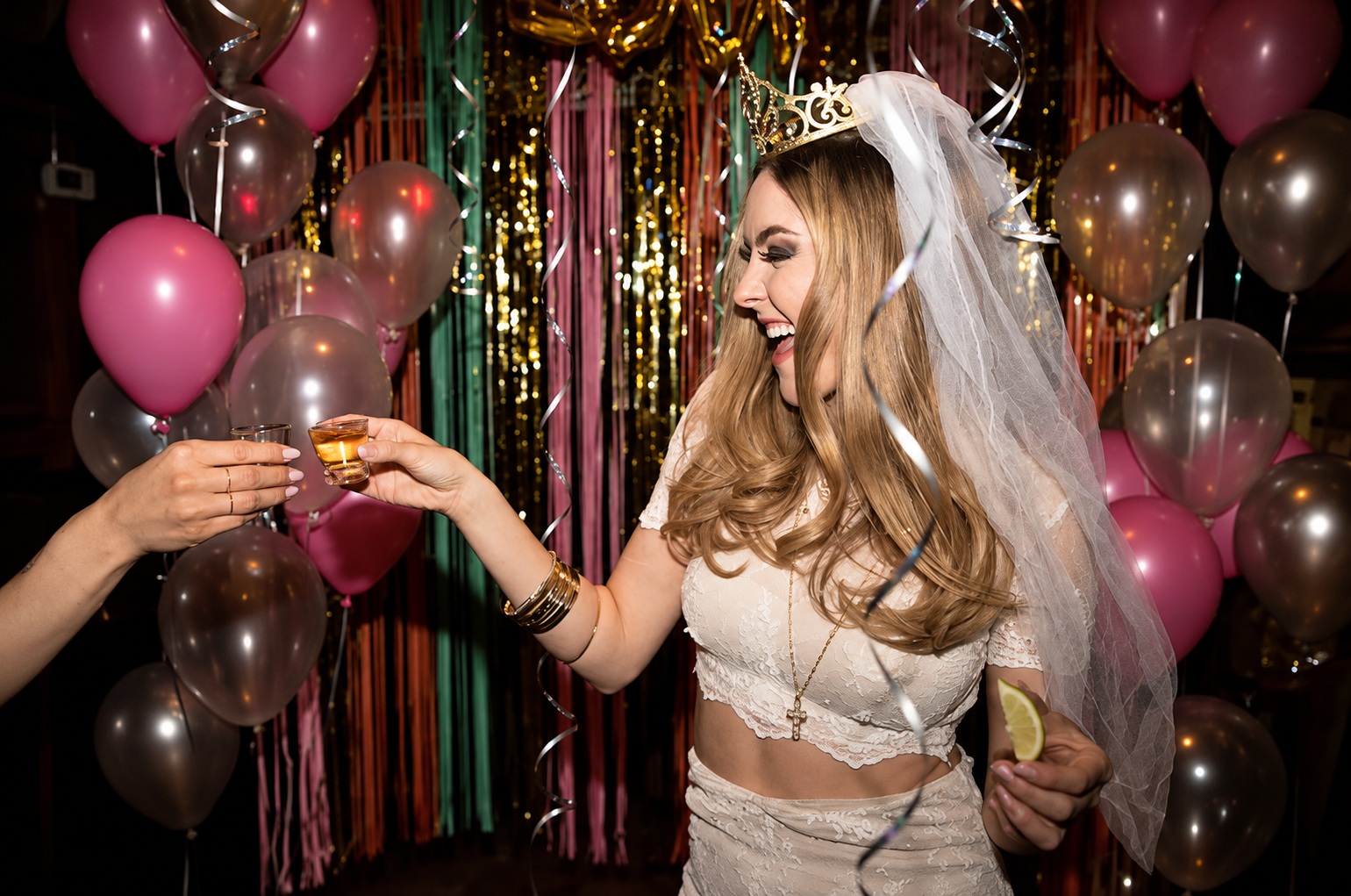 A bride to be in a veil and tiara being handed a drink in front of balloons