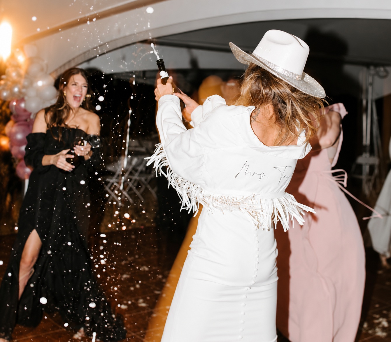 Friends spraying champagne around a bride to be in a white hat