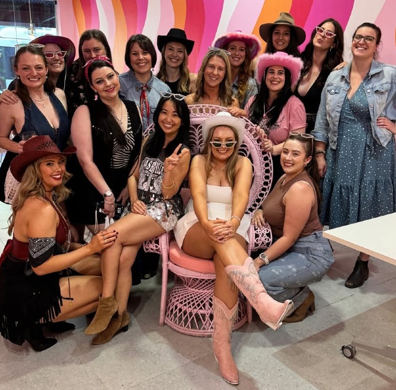 A crew in denim jackets, jeans, cowboy hats and boots around the bride in pink fringed boots on a pink peacock chair