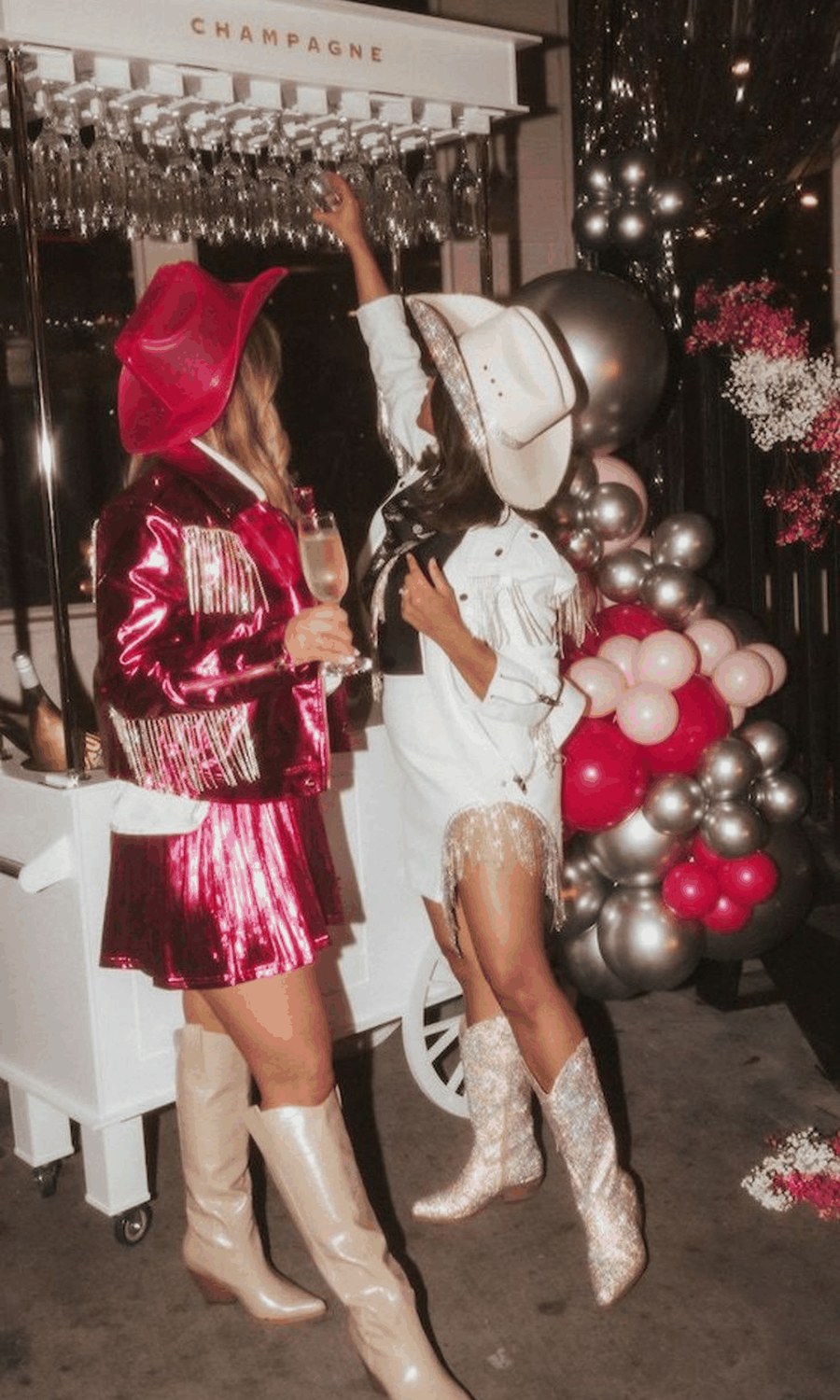 Two friends at a champagne cart under a pink and silver balloon garland, one in a red cowboy hat and hot pink sequin fringe, the other in a white cowboy hat and white fringe, both in cowgirl boots