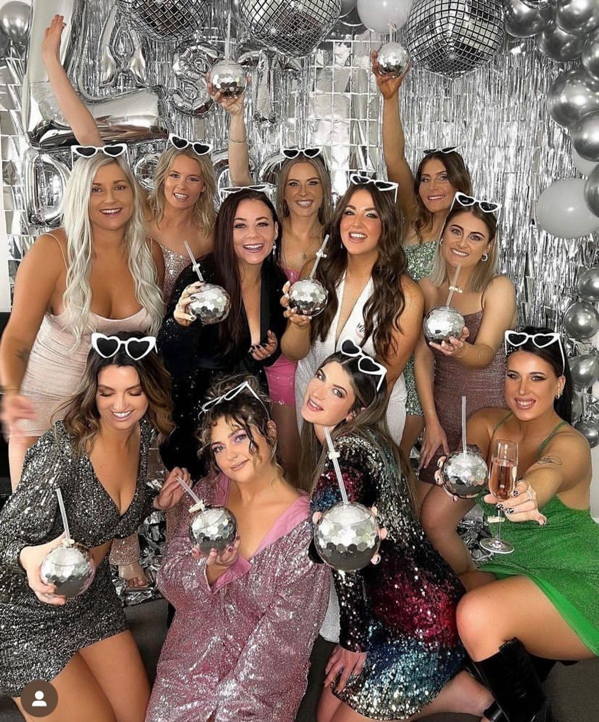 A hens group in sequins and heart sunglasses holding up disco-ball cups in front of a silver balloon wall