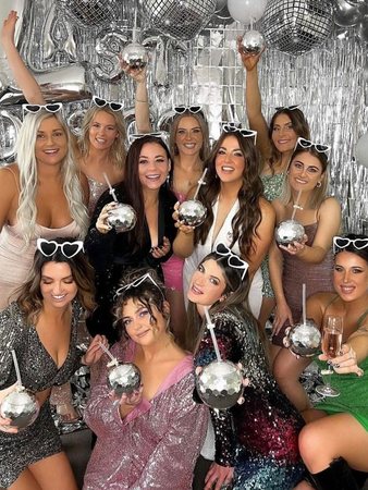 A hens group in sequins holding disco-ball cups in front of a silver balloon wall