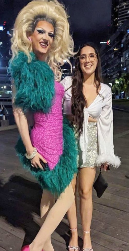Our drag queen in a towering blonde wig and green feather boa posing with a smiling hen on a waterfront boardwalk at night