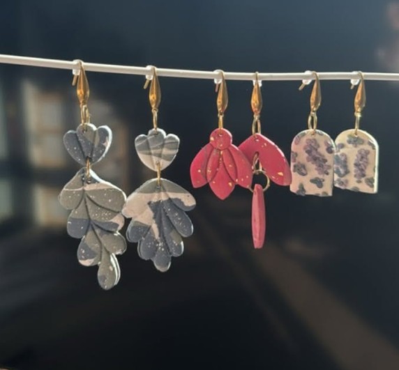Three finished pairs of clay earrings hanging on a line: grey leaves, pink petals and speckled arches