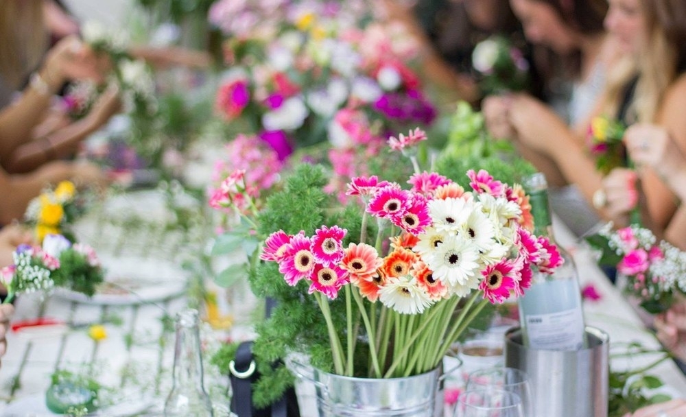 Buckets of bright gerberas and roses on a long table as friends start their flower crowns