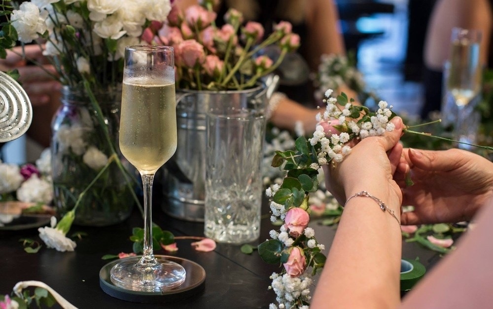 A glass of bubbly beside hands threading a flower crown together at the workshop table
