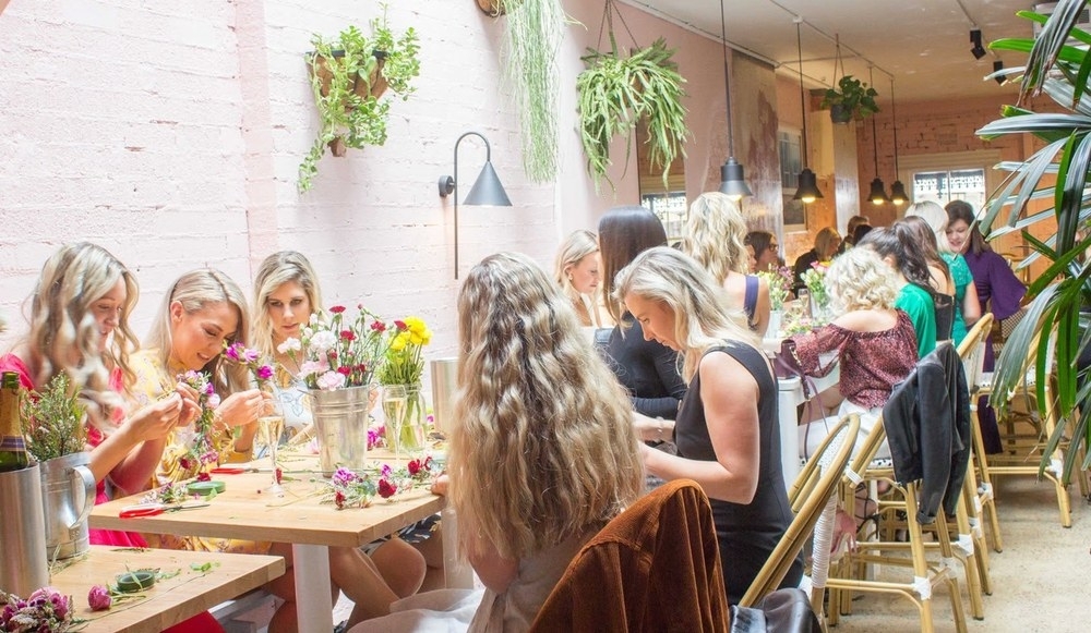 Friends making flower crowns along a long table in a bright room full of flowers