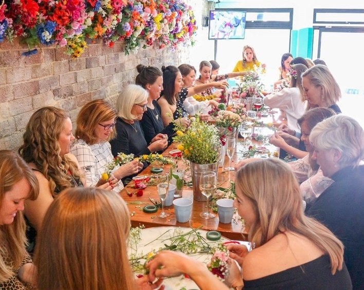 A big group making flower crowns along a long table under a wall of flowers