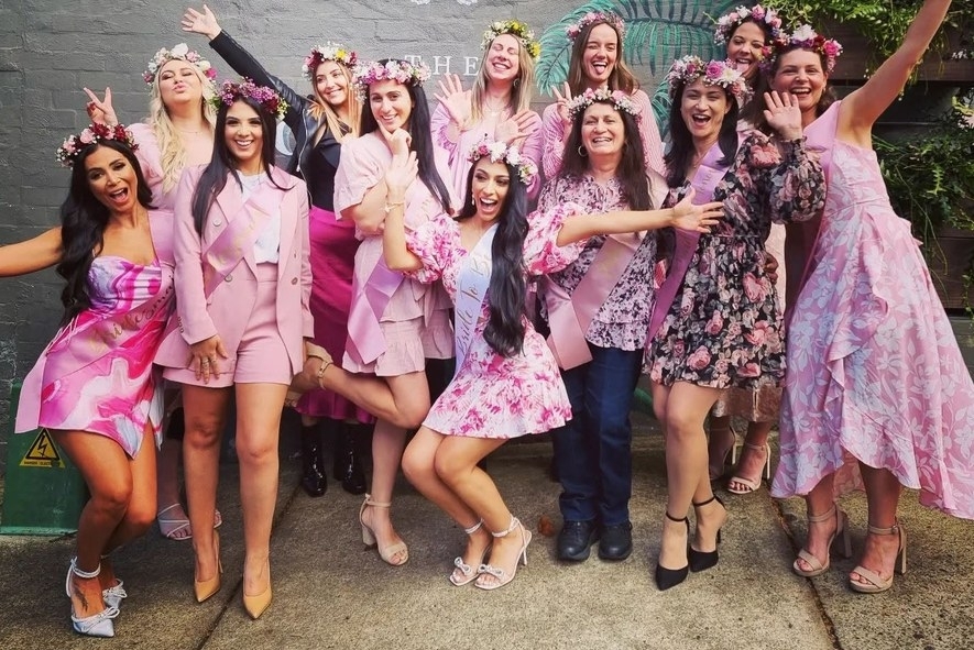 A hens group in pink dresses, flower crowns and sashes cheering together outside