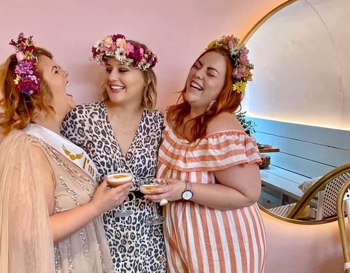 Three friends in bright flower crowns laughing with cocktails in a pink room