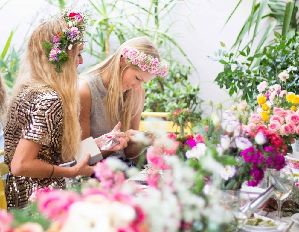 Two friends in flower crowns smiling over the workshop table among buckets of blooms
