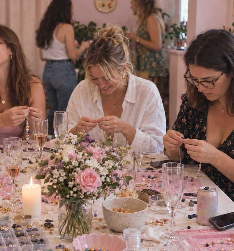 A friend in a white shirt smiles as she threads a bead necklace, behind a vase of pink roses and glasses of rose bubbly