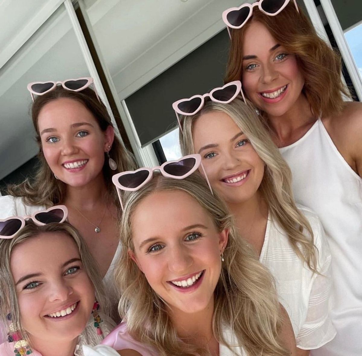 Five friends in white tops and white heart-shaped sunglasses squeezing into a selfie