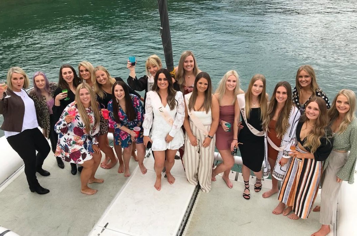 A big hens crew of every age with drinks up on the deck of a catamaran, the bride in white in the middle