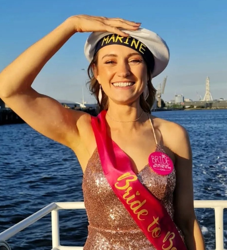 The bride in a sailor hat, sequins and a Bride to Be sash saluting on deck, the bay behind her