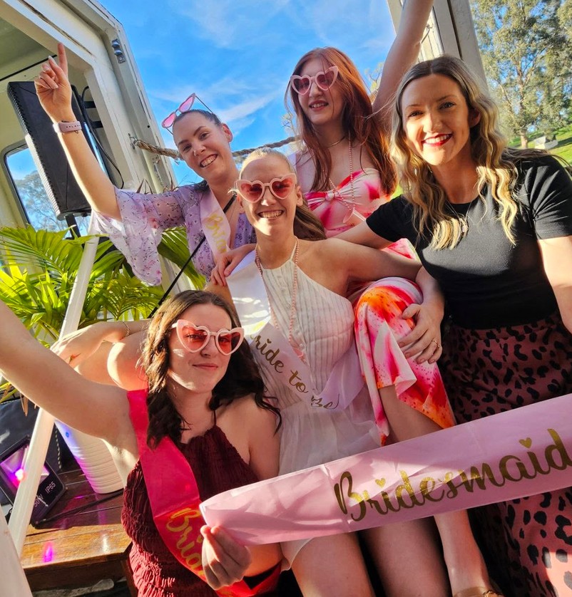 The bride with her bridesmaids in sashes and heart sunglasses on a party boat, arms in the air