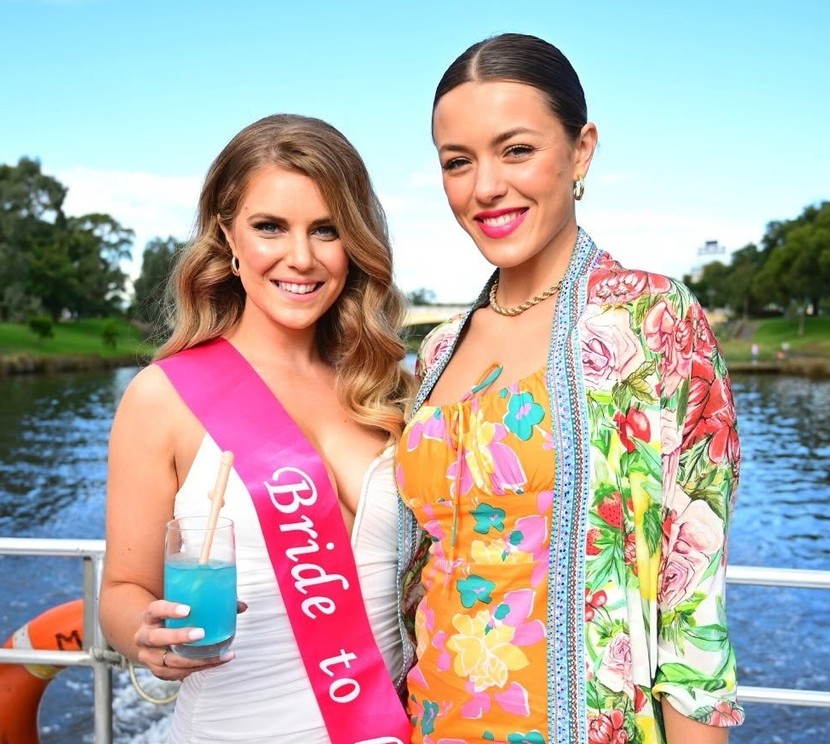 The bride in her pink Bride to be sash with a blue drink beside a friend in florals, the river behind them